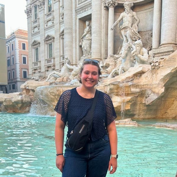 Person standing in front of the Trevi Fountain in Rome wearing a black top, blue jeans, and white sneakers with a crossbody bag.