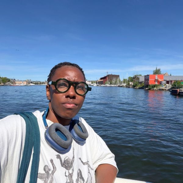 Person wearing a white shirt and headphones around their neck stands by a waterfront with boats and buildings under a clear blue sky.