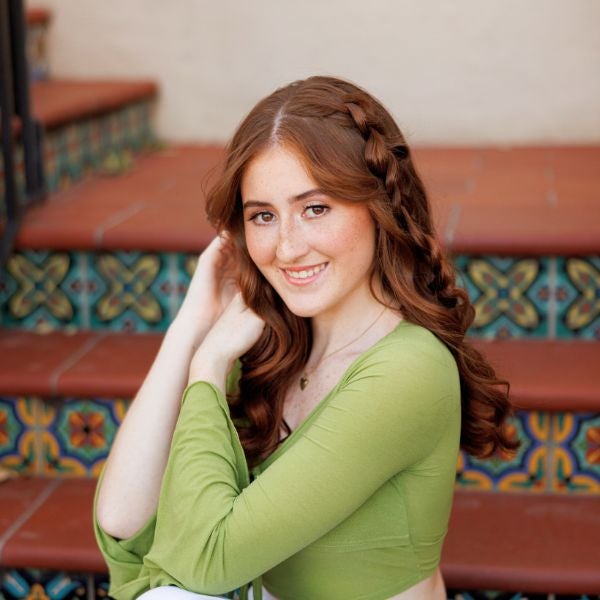 Woman with long red hair styled in a braid, wearing a green top, sitting on colorful tiled stairs indoors.
