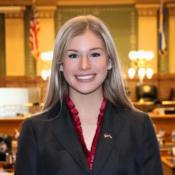Woman in formal attire standing in a government chamber with American flags and wooden decor in the background.