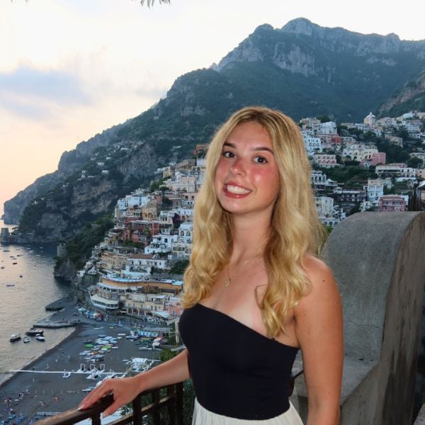 Woman with long blonde hair in a black strapless top and white skirt stands by a railing overlooking the a coastal town at sunset.