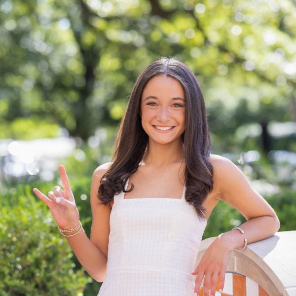 Young woman in a white sleeveless dress stands outdoors, making a hand gesture with her left hand while leaning on a tiled wall.