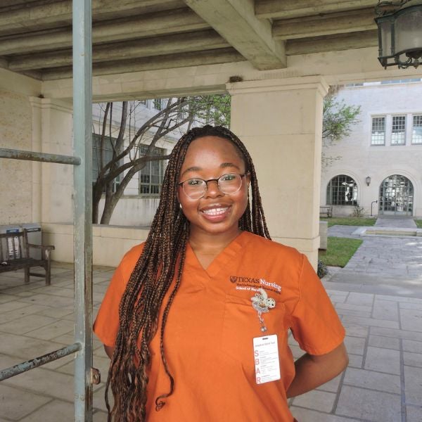 Person wearing an orange Texas Nursing uniform standing outdoors under a covered walkway with long braided hair.