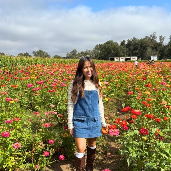 Young woman in a denim dress and boots walking through a vibrant flower field under a partly cloudy sky.