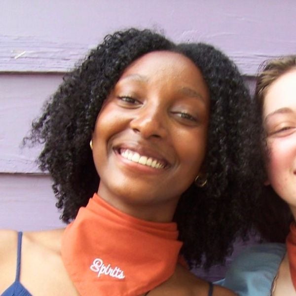 Person with curly hair wearing an orange neck gaiter with the word 'Splitts' embroidered, against a light purple wooden wall.