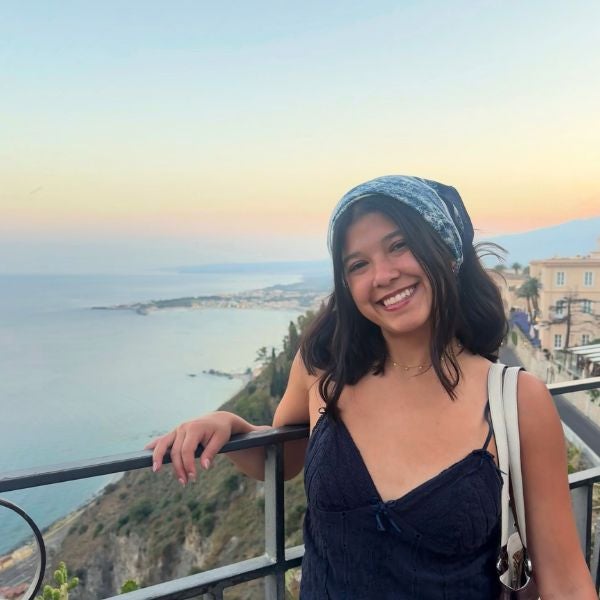Woman in a black dress and headscarf leaning on a balcony railing overlooking a coastal cliff and calm sea at sunset.