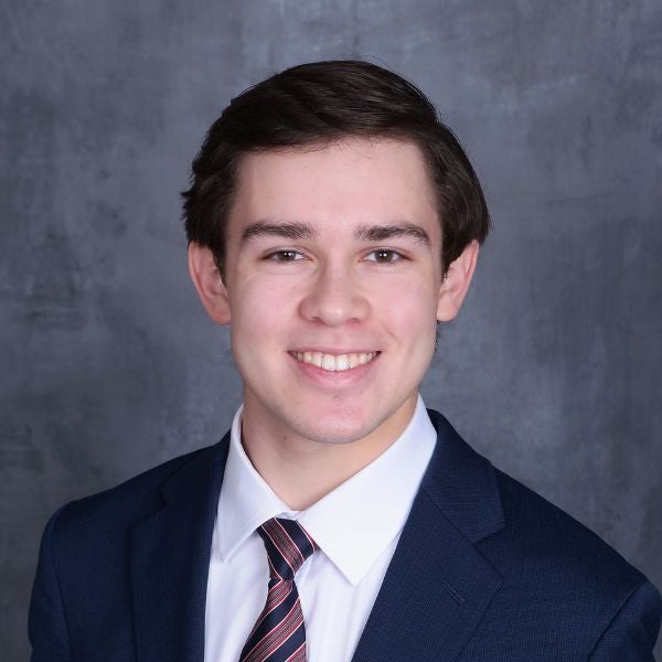 Young man in a navy blue suit and striped tie posing against a gray textured backdrop.