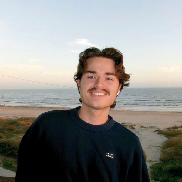 Person wearing a dark Alo sweatshirt standing near a beach with calm ocean waves and a clear sky at sunset.