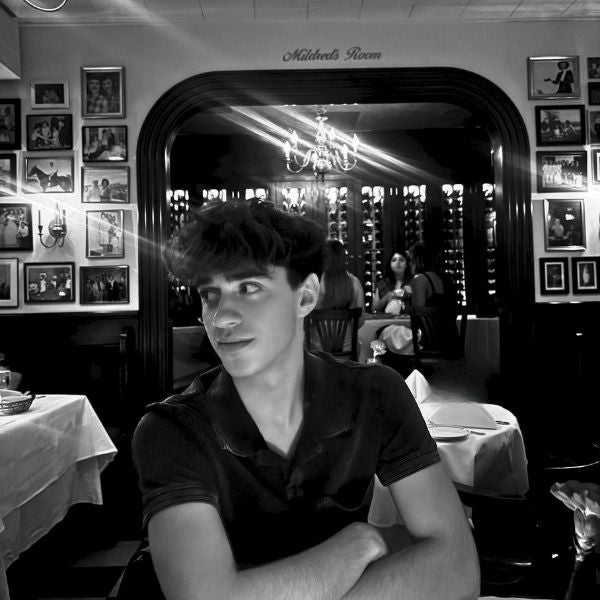 Black and white photo of a young man sitting at a table in a vintage restaurant room with framed pictures on the walls.