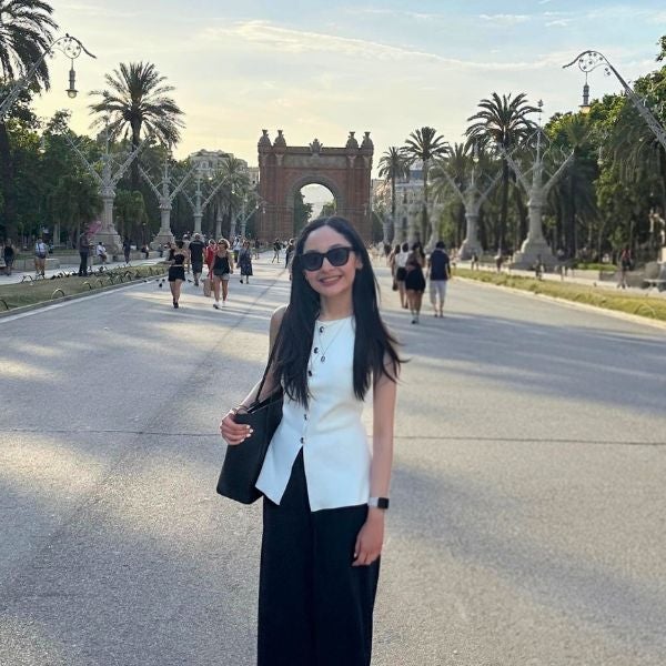 Woman in white sleeveless top and black pants standing on a wide pedestrian street with the Arc de Triomf in Barcelona in the background.