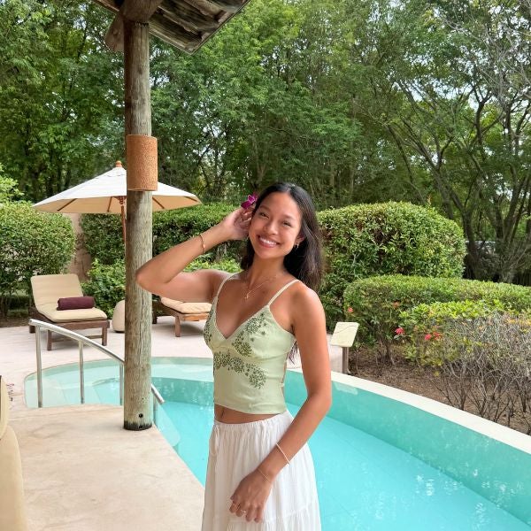 Woman in a light green camisole and white skirt posing by a curved pool surrounded by lush greenery and lounge chairs.