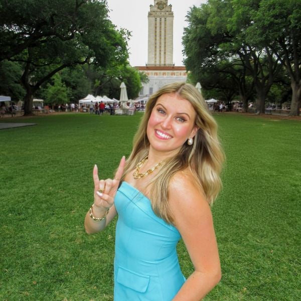 Woman in a light blue strapless dress making the 'Hook 'em Horns' hand sign on a green lawn with a tall clock tower in the background.