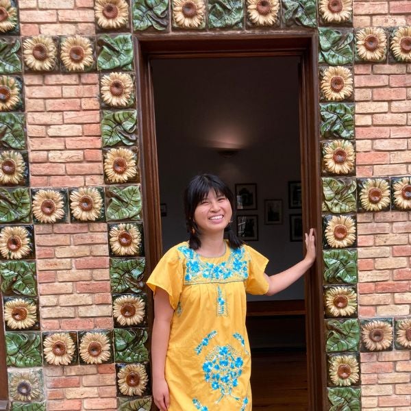 Person in a yellow dress with blue floral embroidery stands in a doorway framed by decorative sunflower tiles on a brick wall.
