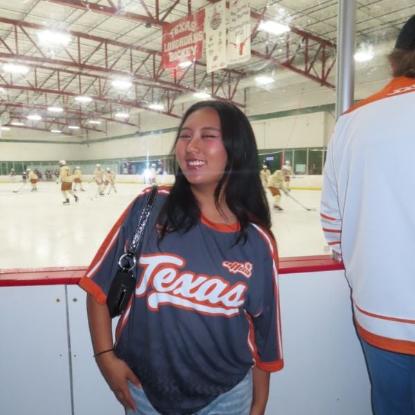 Woman wearing a Texas sports jersey stands inside an ice hockey rink with players practicing in the background.