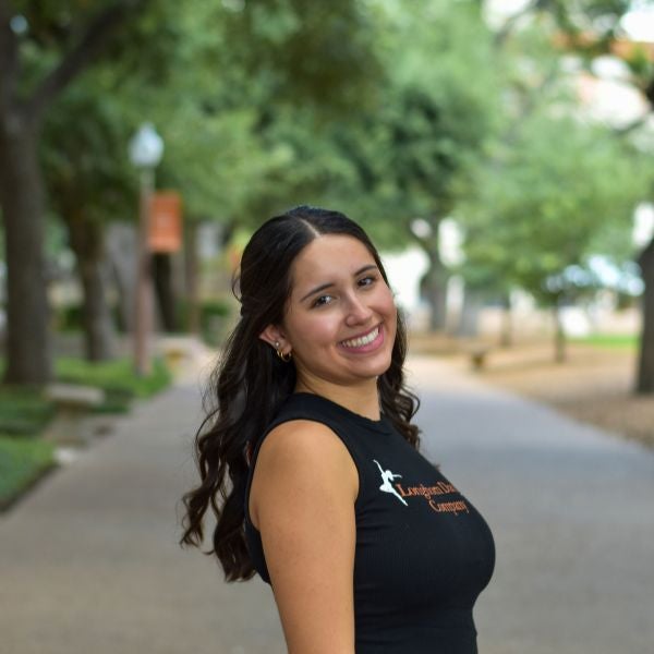 Young woman with long dark hair wearing a black sleeveless shirt standing on a tree-lined pathway outdoors.
