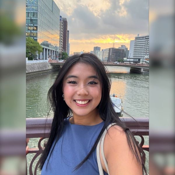 Young woman smiling on a bridge over a river with city buildings and a sunset sky in the background.