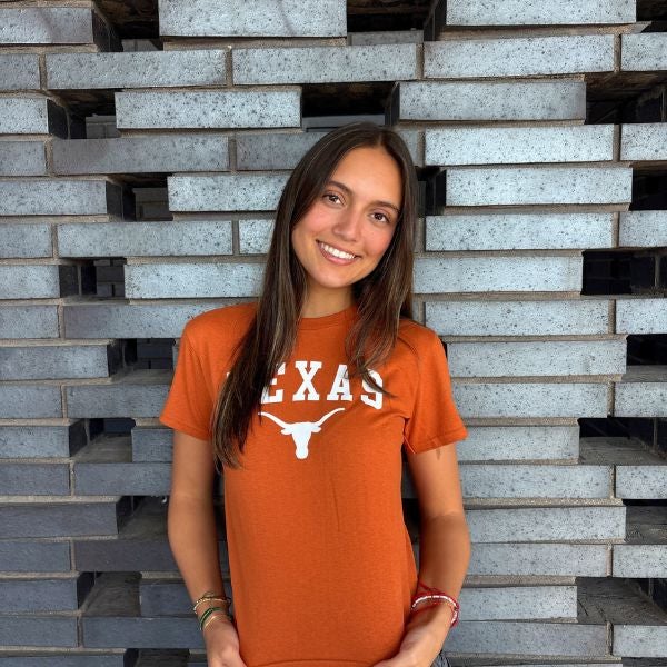 Young woman smiling in front of a modern gray brick wall wearing a burnt orange Texas Longhorns t-shirt.