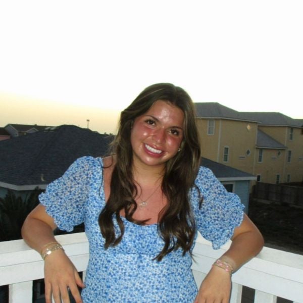 Young woman with long brown hair wearing a blue floral dress smiles while leaning on a white balcony railing at sunset.