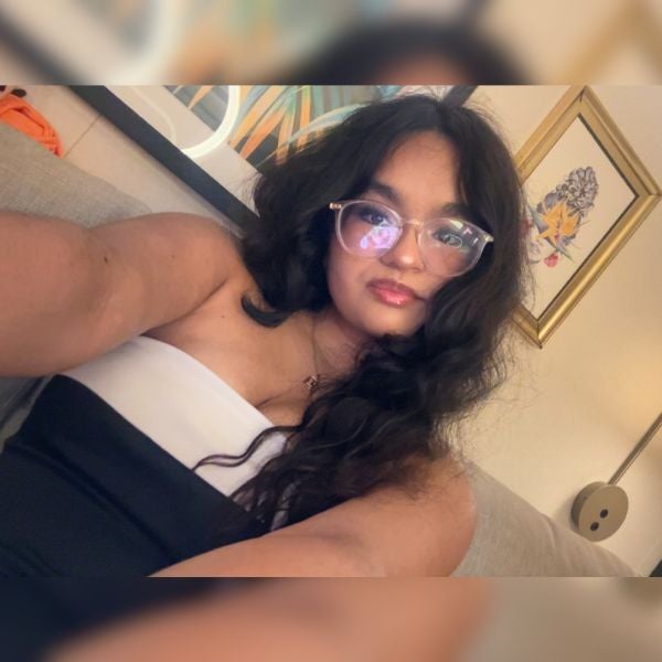 Young woman with long dark hair and glasses wearing a strapless black and white top sitting on a couch indoors.