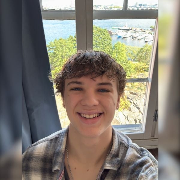 Smiling young man with short curly hair wearing a plaid shirt, standing indoors by a window overlooking a marina and green trees.
