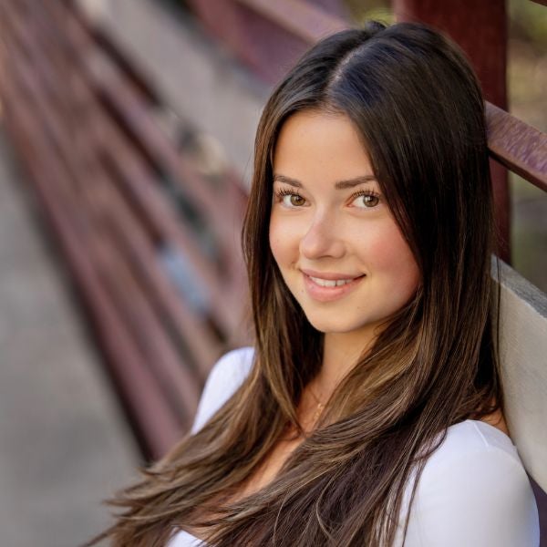 Young woman with long brown hair and a white top smiling softly while leaning against a wooden railing outdoors.
