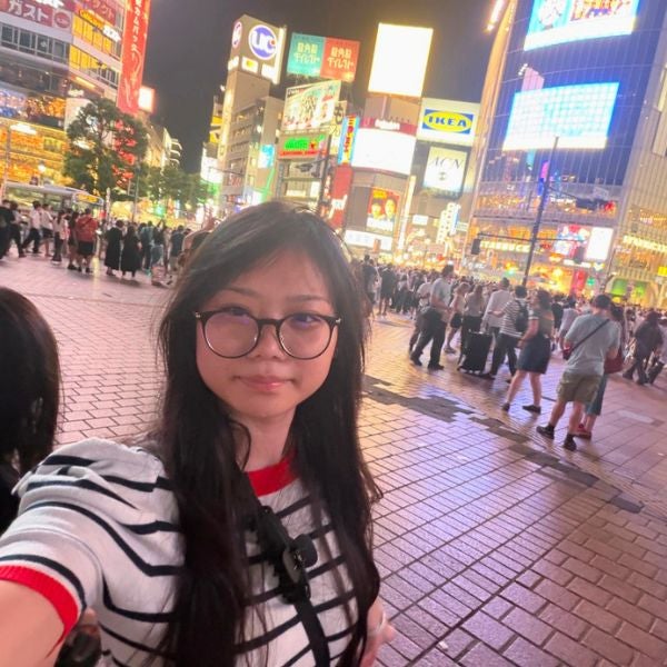 Young woman with glasses taking a selfie at a busy, brightly lit urban square at night filled with people and illuminated billboards.