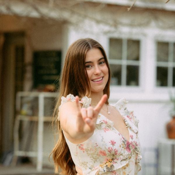 Young woman with long brown hair smiling and making a hand gesture in a floral dress outdoors.