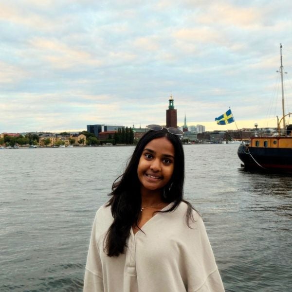 Young woman smiling by a waterfront with a Swedish flag and cityscape in the background under a cloudy sky.