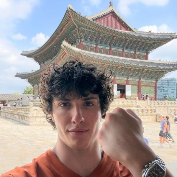 Young man with curly hair poses with a fist in front of a traditional Korean palace under a blue sky.