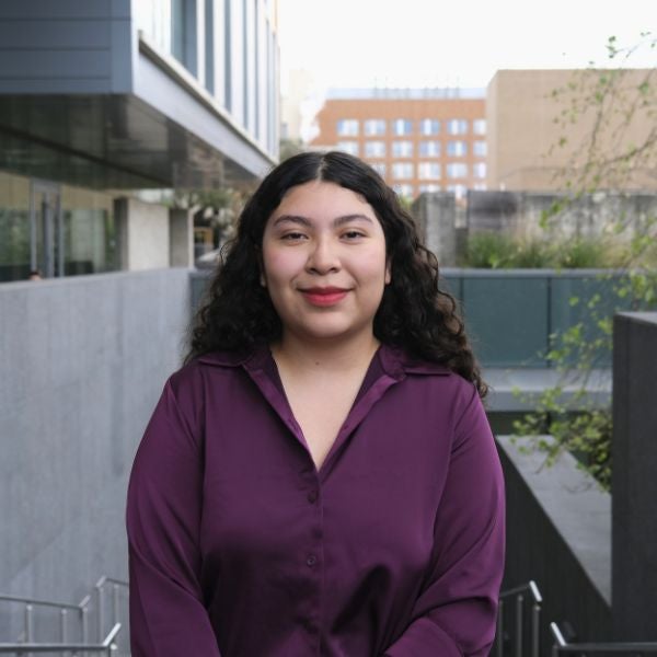 Young woman with curly dark hair wearing a purple blouse stands outdoors in an urban setting with buildings in the background.