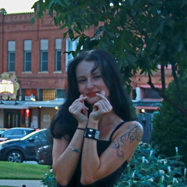 Young woman with dark hair and a tattoo on her arm making a heart gesture with her hands outdoors near a tree and parked cars.