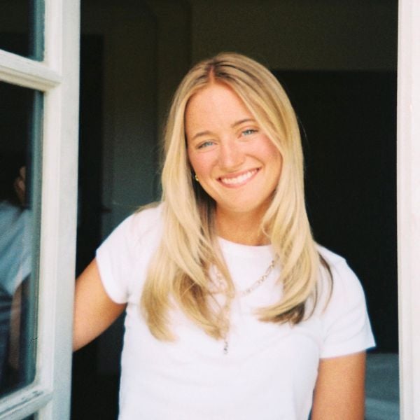 Smiling young woman with long blonde hair wearing a white t-shirt standing in a doorway.