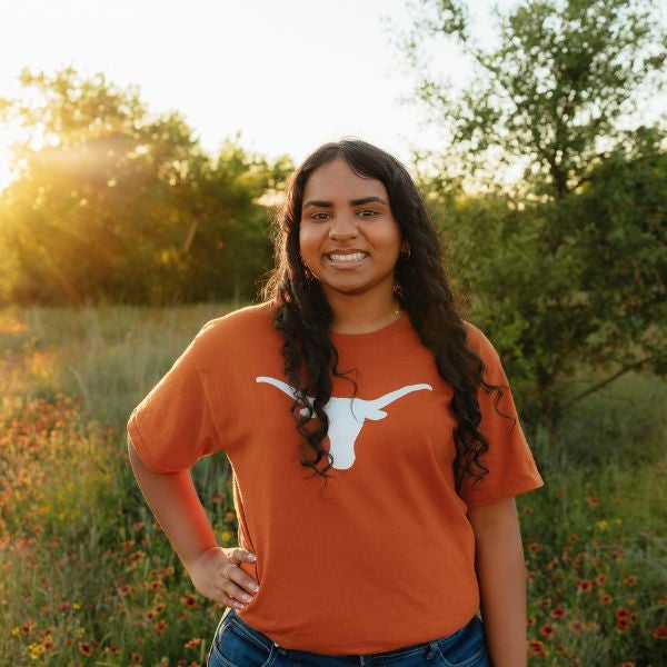 Young woman with long curly hair wearing an orange Texas Longhorns t-shirt stands smiling in a sunlit field of wildflowers.
