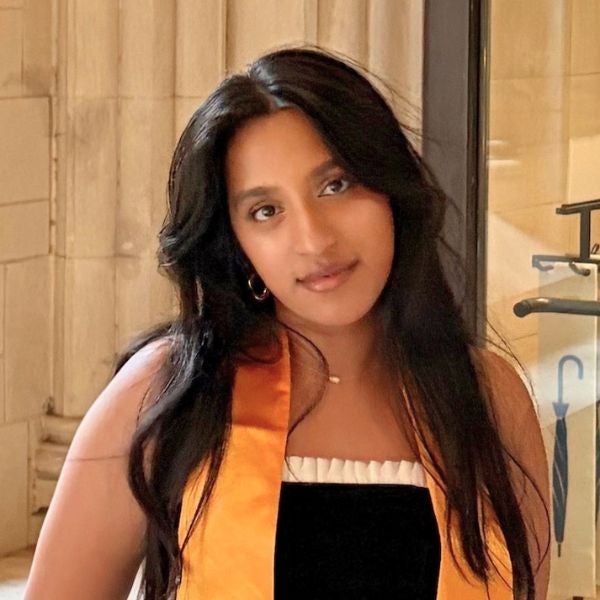 Young woman with long dark hair wearing an orange graduation stole and black dress, standing indoors near a glass door.