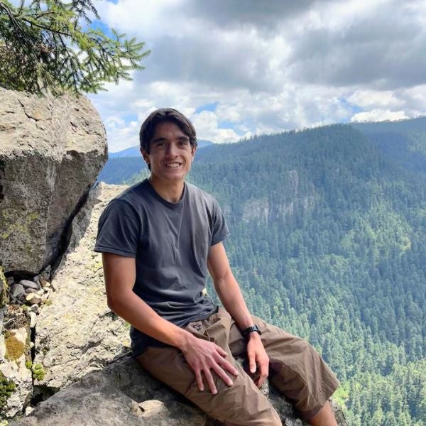 Young man sitting on a rocky ledge overlooking a vast forested mountain landscape under a partly cloudy sky.