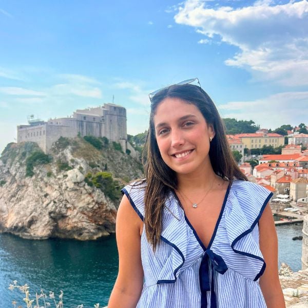 Young woman smiling in a blue and white ruffled dress with a historic stone fortress and coastal town in the background under a partly cloudy sky.