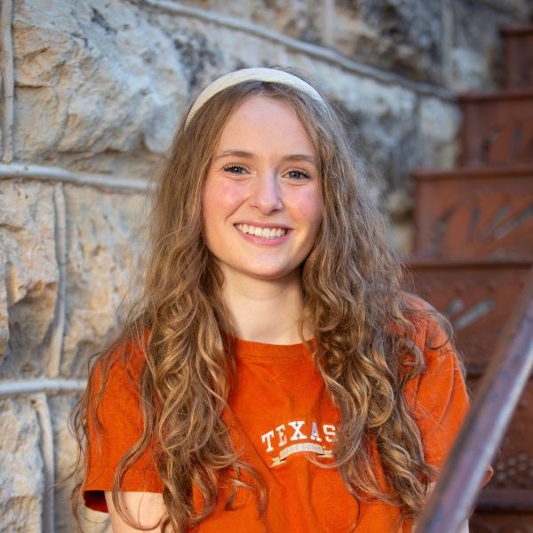 Young woman with long curly hair wearing an orange Texas t-shirt smiles while sitting by a stone wall and metal staircase.