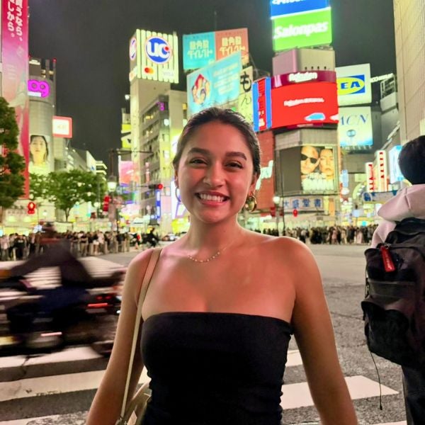 Smiling woman in a black strapless top standing at a busy, brightly lit urban intersection at night.