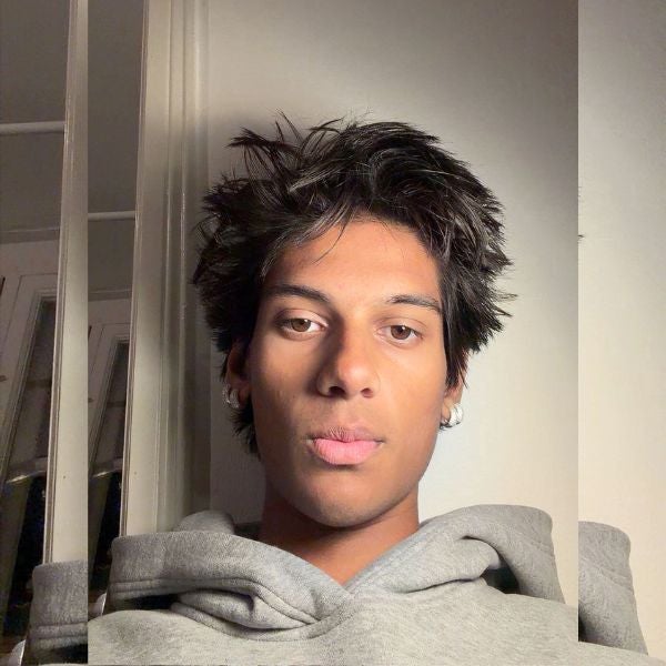Young man with tousled black hair wearing a gray hoodie, looking directly at the camera against a plain background.