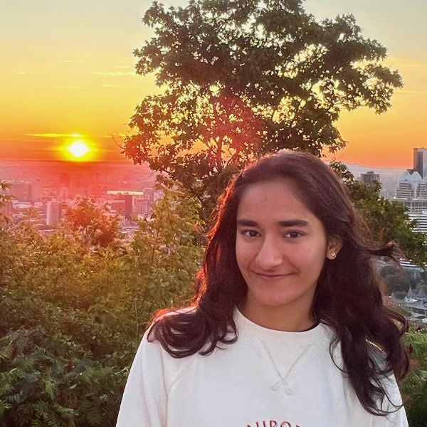 Young woman with long dark hair smiles softly in front of a vibrant sunset over a cityscape and lush greenery.